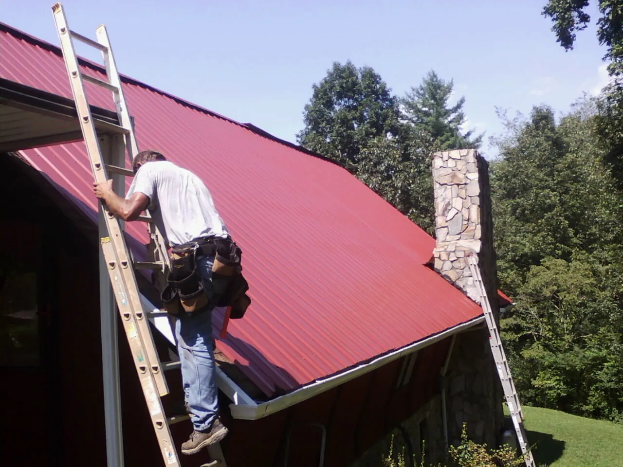 Roofer on a ladder beside a red metal roof and stone chimney — Soffit & Fascia Repair by Roofing Brothers in Saint Ansgar